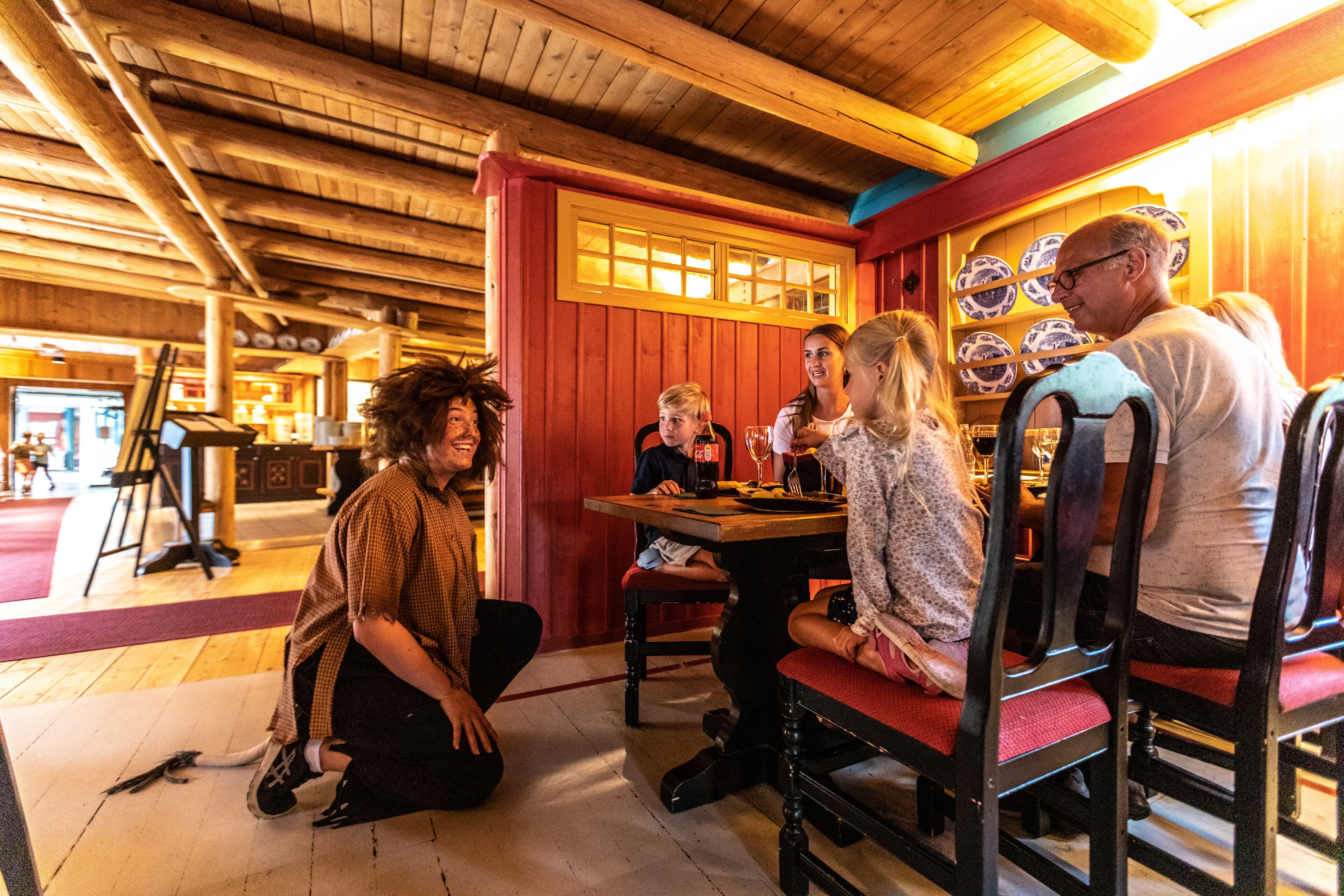Un groupe de personnes est assis autour d’une table en bois, dans un intérieur rustique avec des poutres apparentes au plafond et un mur rouge agrémenté d’une fenêtre.