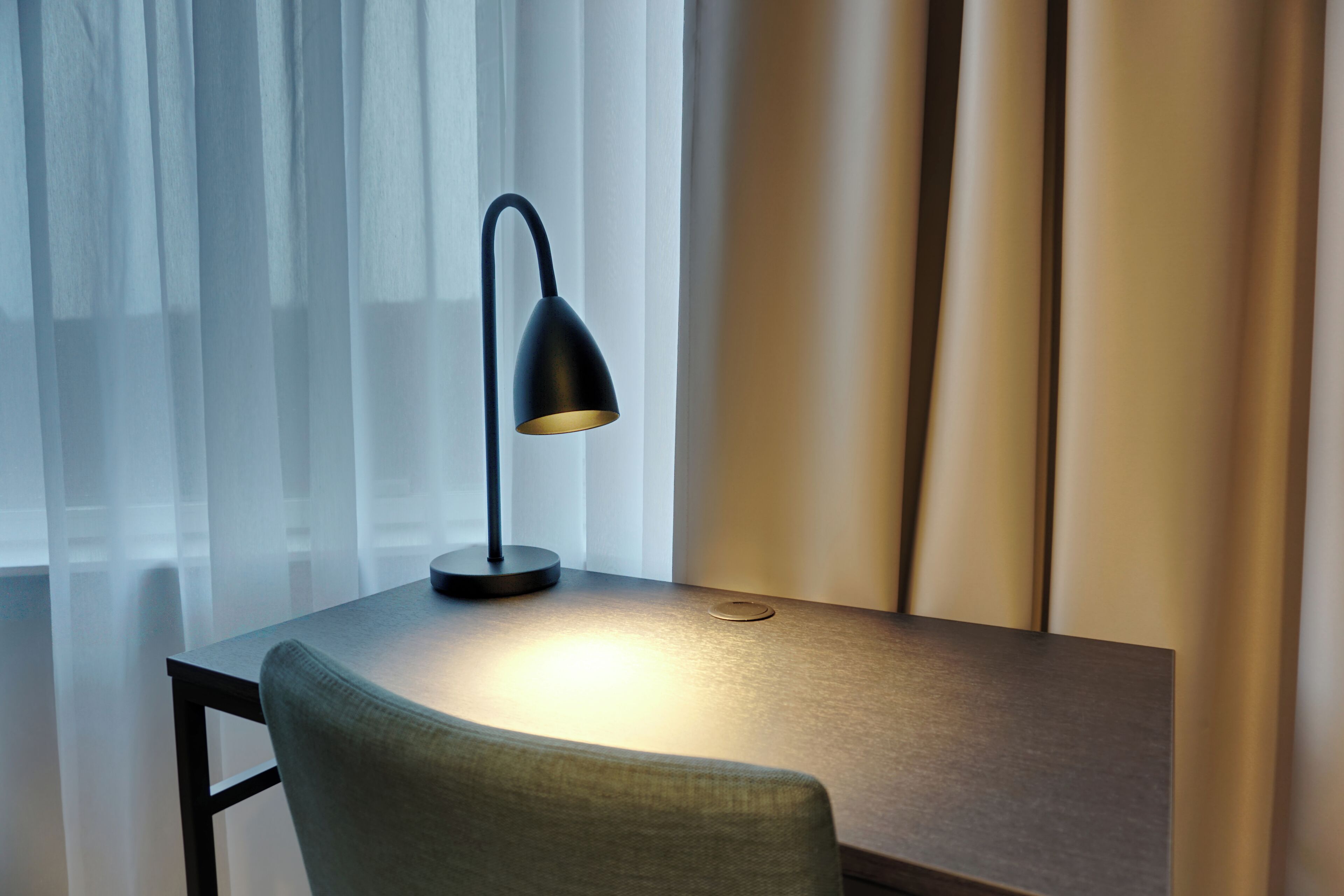Eine moderne Schreibtischlampe auf einem Tisch in der Nähe eines Fensters mit durchsichtigen Vorhängen.