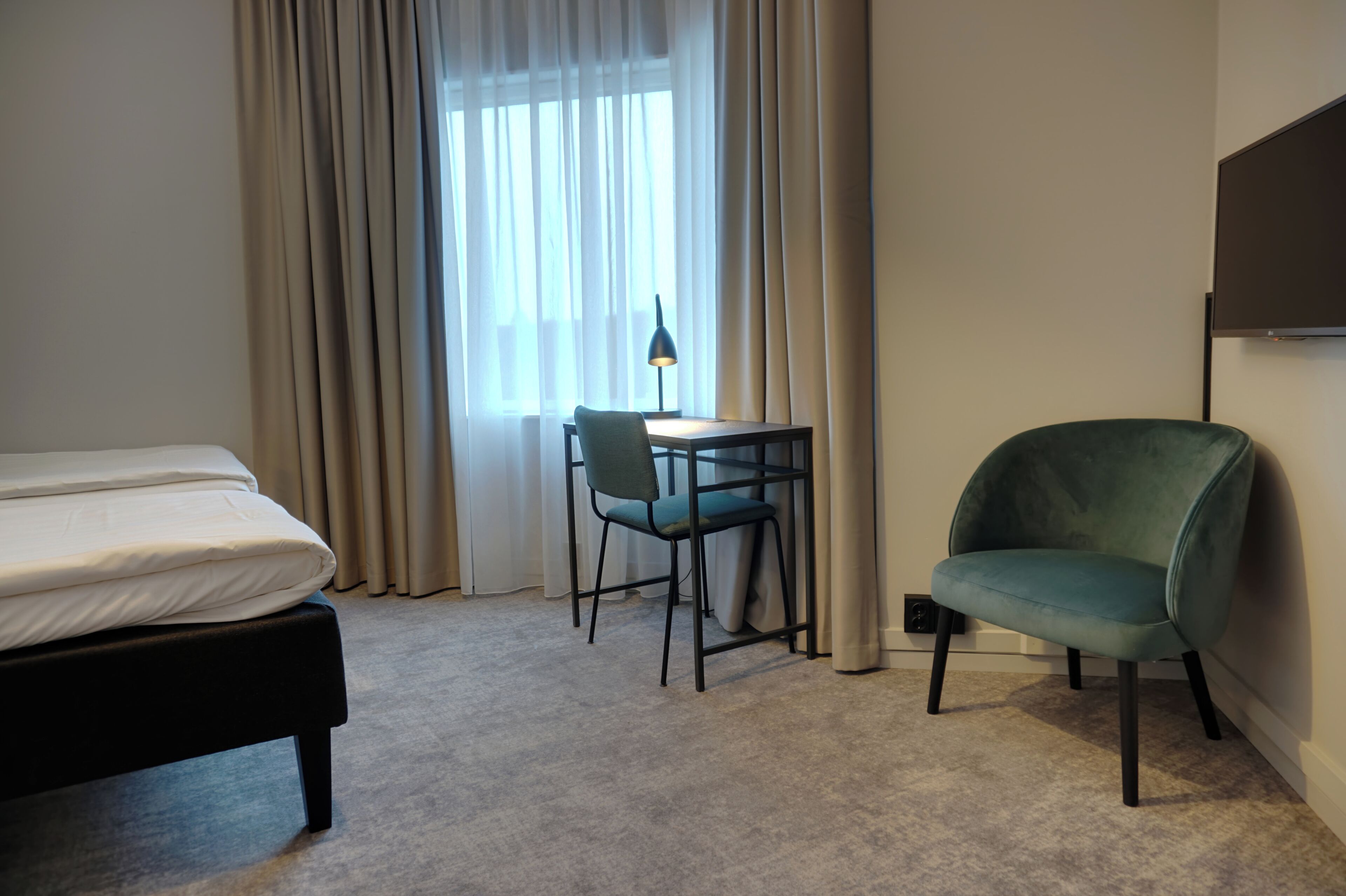 A hotel room with a bed, a desk with a chair, a TV, and a window with curtains.