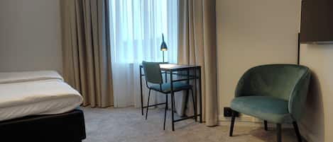 A hotel room with a bed, a desk with a chair, a TV, and a window with curtains.