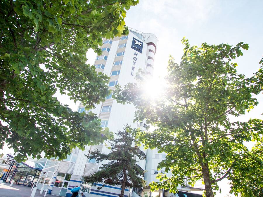 Een modern hotelgebouw met een opvallend 'First Hotel' bord, omringd door weelderige groene bomen.
