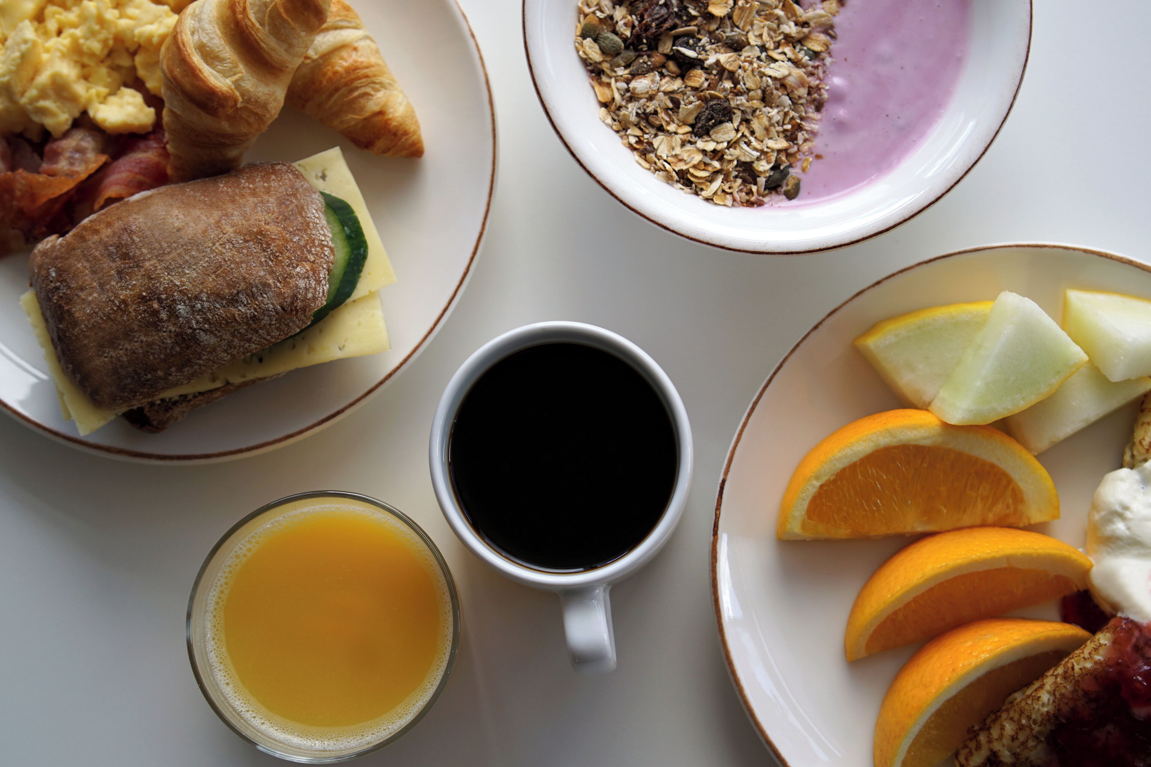 A breakfast table with croissants, scrambled eggs, bacon, a sandwich, a bowl of granola, a cup of coffee, a glass of orange juice, and sliced oranges.
