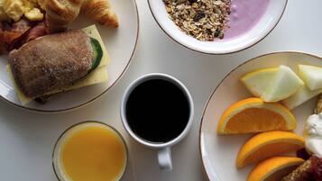 Ein Frühstückstisch mit Croissants, Rührei, Speck, einem Sandwich, einer Schüssel Granola, einer Tasse Kaffee, einem Glas Orangensaft und Orangenscheiben.