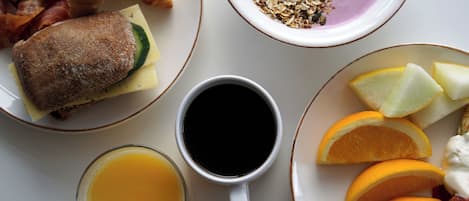 Una mesa de desayuno con croissants, huevos revueltos, tocino, un sándwich, un tazón de granola, una taza de café, un vaso de jugo de naranja y rodajas de naranja.