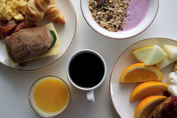 Una mesa de desayuno con croissants, huevos revueltos, tocino, un sándwich, un tazón de granola, una taza de café, un vaso de jugo de naranja y rodajas de naranja.