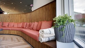 A modern interior with a curved wooden bench, a large grey vase with green plants, and a view through a window.