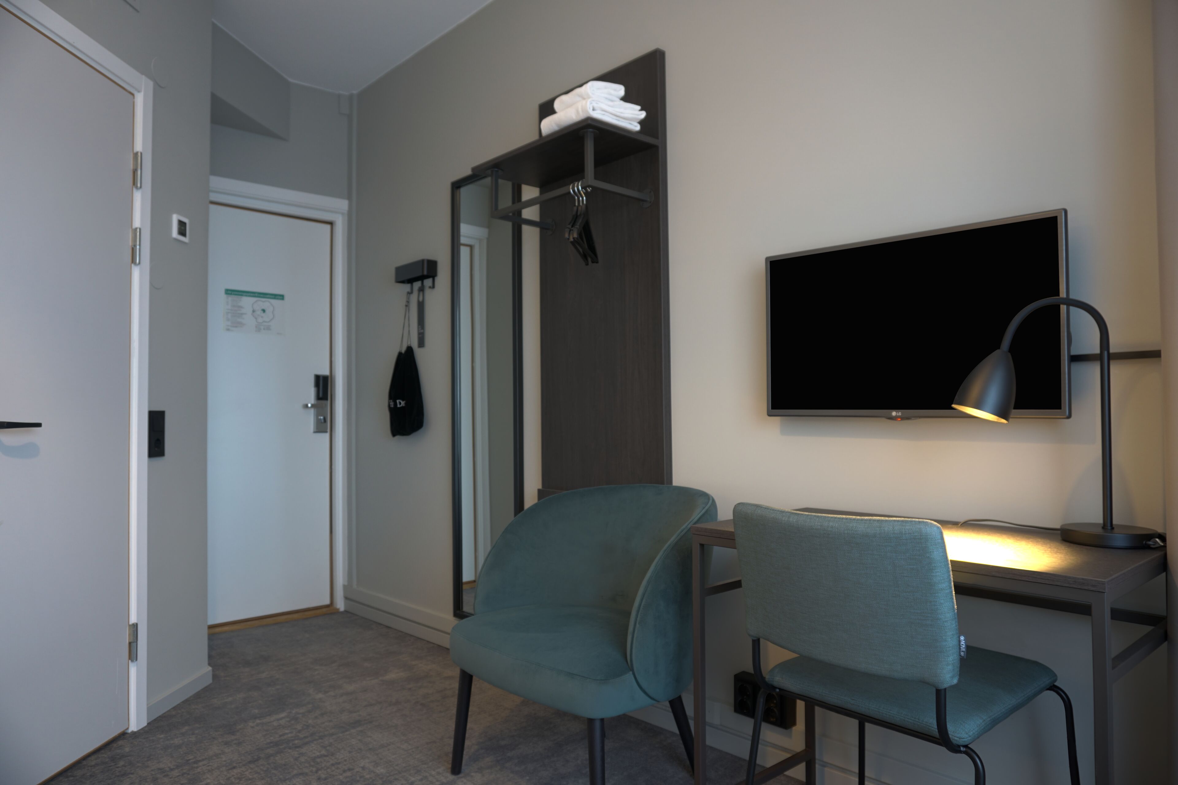 A hotel room with a desk, two chairs, a TV, a lamp, a coat rack with folded clothes, and a door with a sign.
