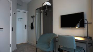 Habitación de hotel con escritorio, dos sillas, televisor, lámpara, perchero con ropa doblada y una puerta con un cartel.