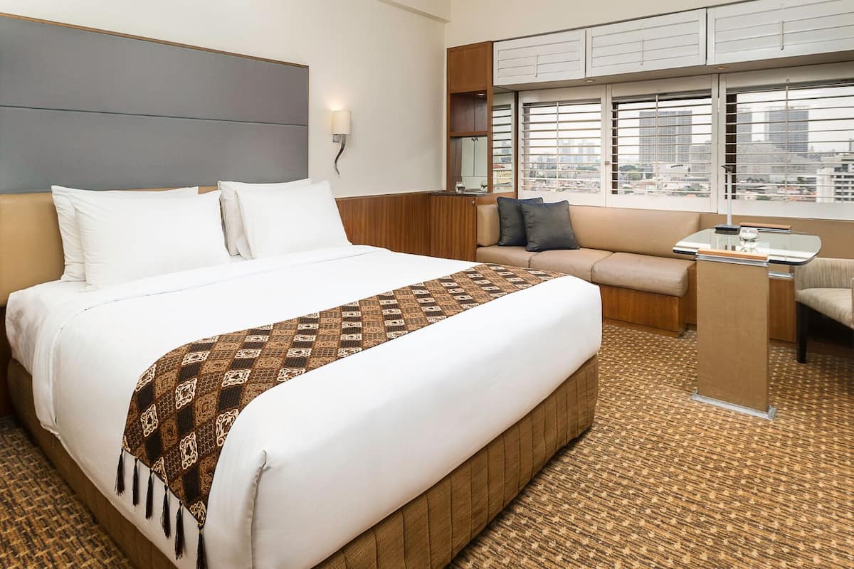 Ein Hotelzimmer mit einem großen Bett, einem kleinen Schreibtisch, einer Sitzecke und Blick auf die Stadt durch große Fenster.