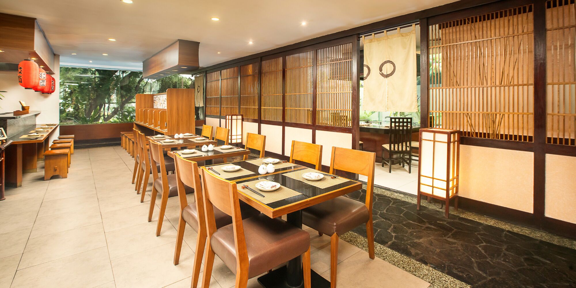 A restaurant with multiple tables and chairs, a view of greenery outside, and traditional decor elements like wooden partitions and lanterns.