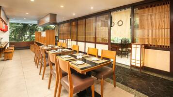 A restaurant with multiple tables and chairs, a view of greenery outside, and traditional decor elements like wooden partitions and lanterns.