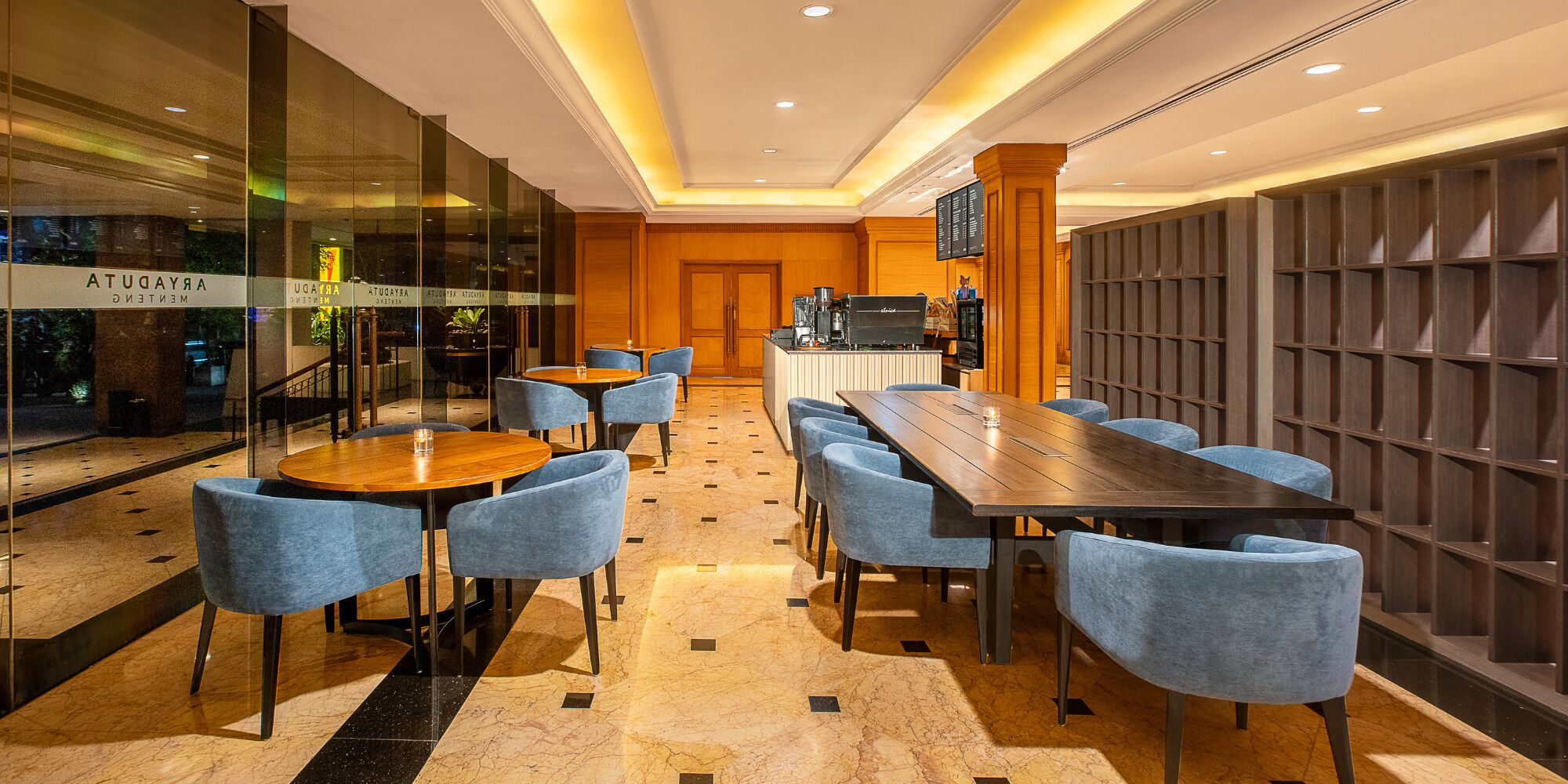 A modern hotel lobby with a long wooden table and blue chairs, a bar area, and a glass wall with signage.