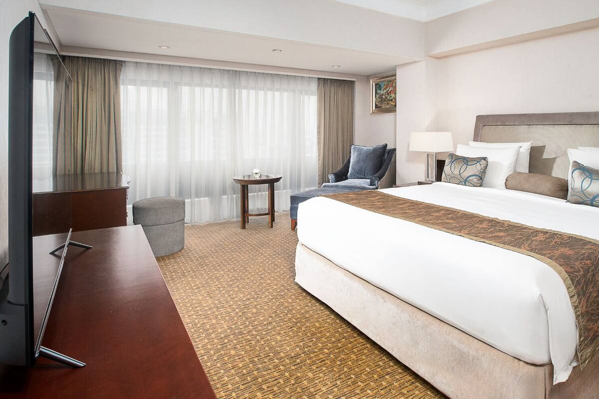 Ein Hotelzimmer mit einem großen Bett, einem Schreibtisch, einem Stuhl, einem kleinen Tisch und einem Fenster mit Vorhängen.