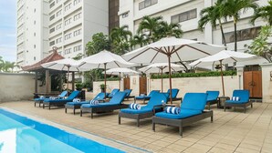 Un área junto a la piscina con sillones de descanso y sombrillas, un suelo de baldosas y un edificio al fondo.