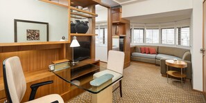 A modern hotel room with a glass desk, a sofa, a small round table, and a large window with blinds.