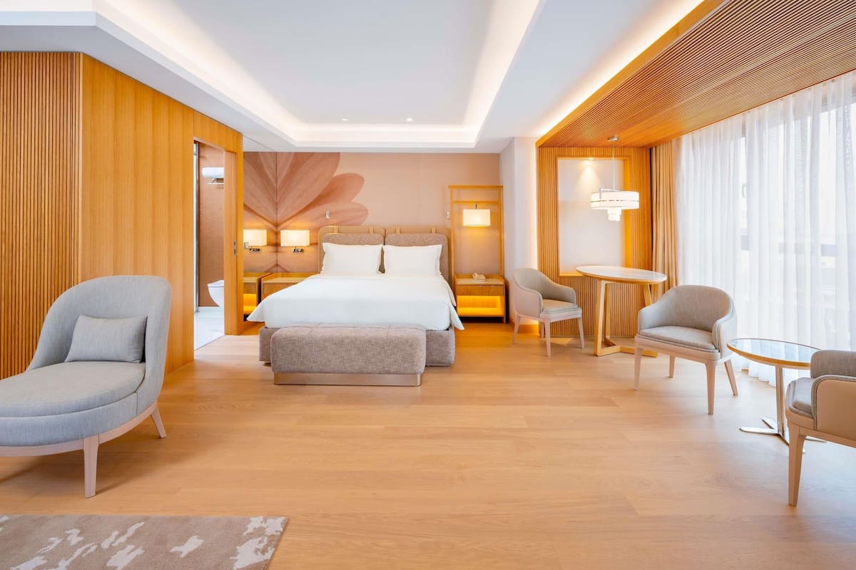 A modern hotel room with a large bed, two armchairs, a small table, and a view of the city through large windows.
