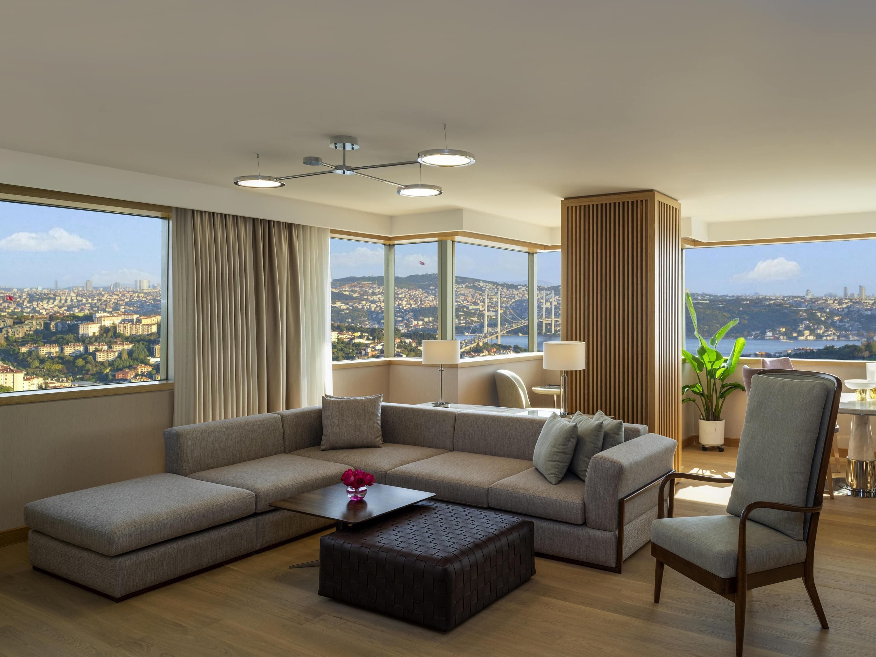 Ein modernes Wohnzimmer mit einem Ecksofa, einem Couchtisch und Blick auf eine Stadtlandschaft durch große Fenster.