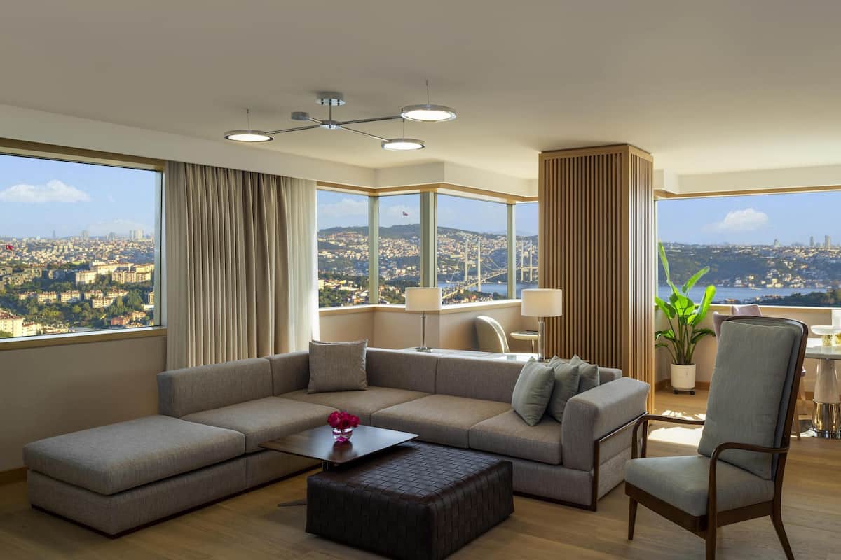 Ein modernes Wohnzimmer mit einem Ecksofa, einem Couchtisch und Blick auf eine Stadtlandschaft durch große Fenster.