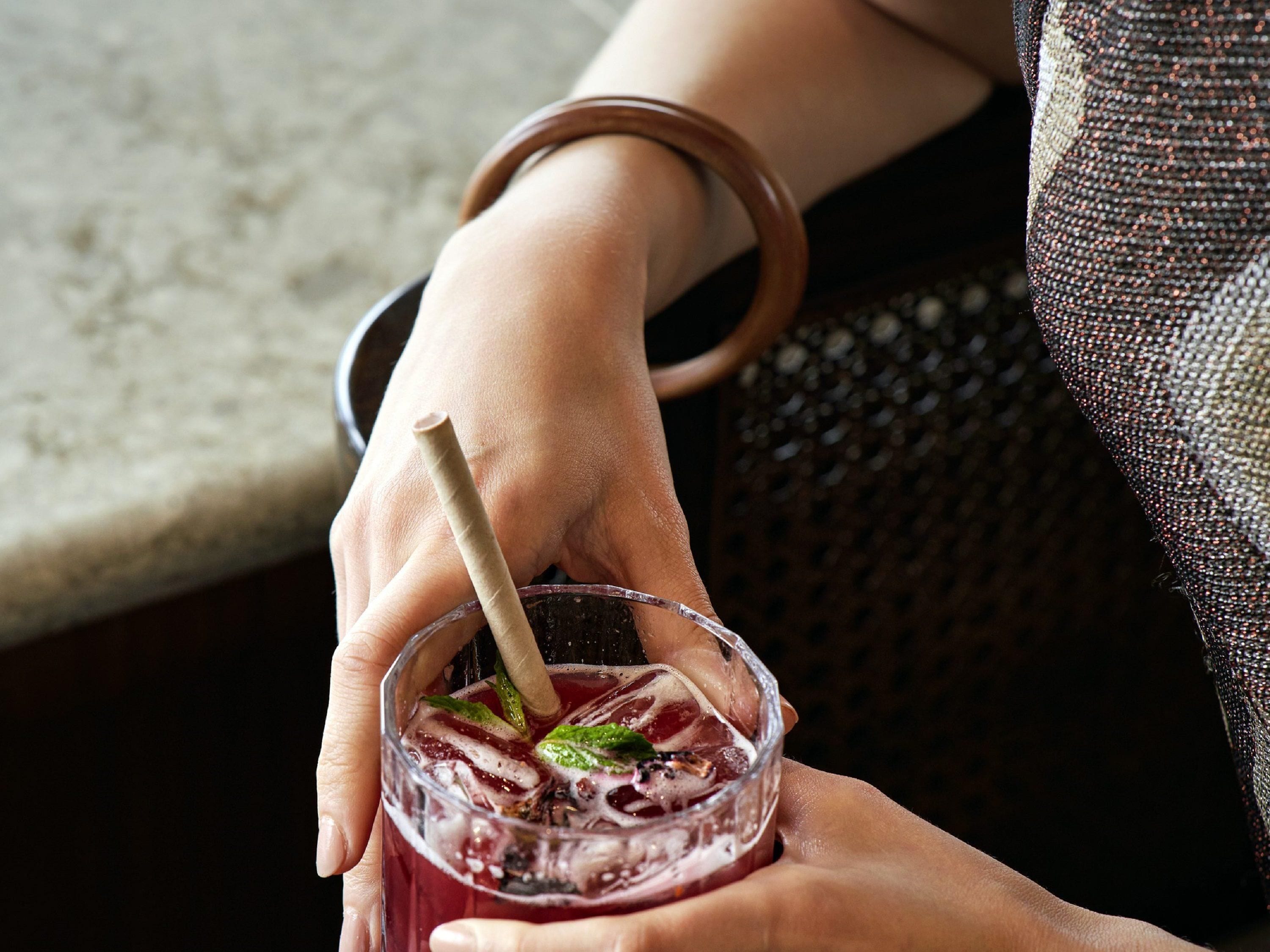 A close-up of a hand holding a glass with a straw, containing a beverage with ice and mint leaves.