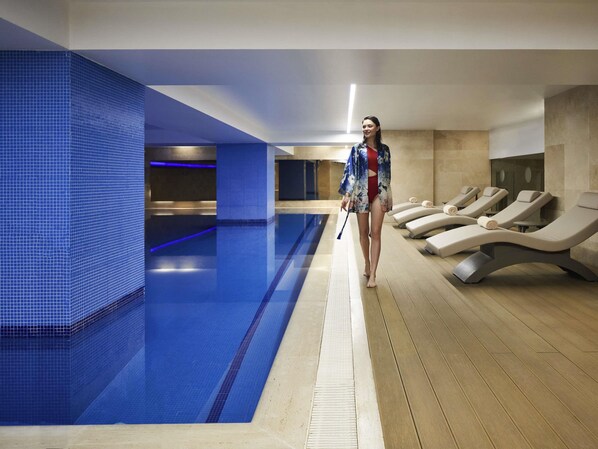 Una piscina con baldosas azules, un área de descanso con tumbonas y una persona caminando con una toalla.