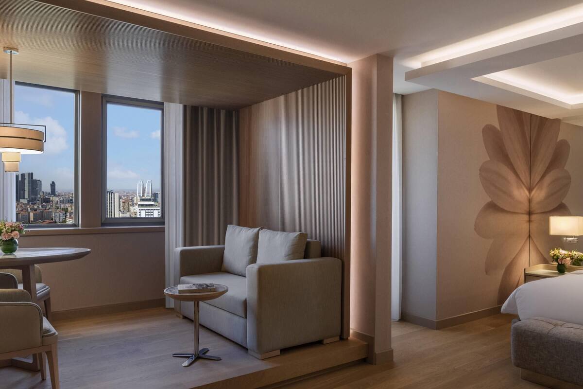 A modern hotel room with a sofa, a small round table, a bed, and a large window offering a city view.