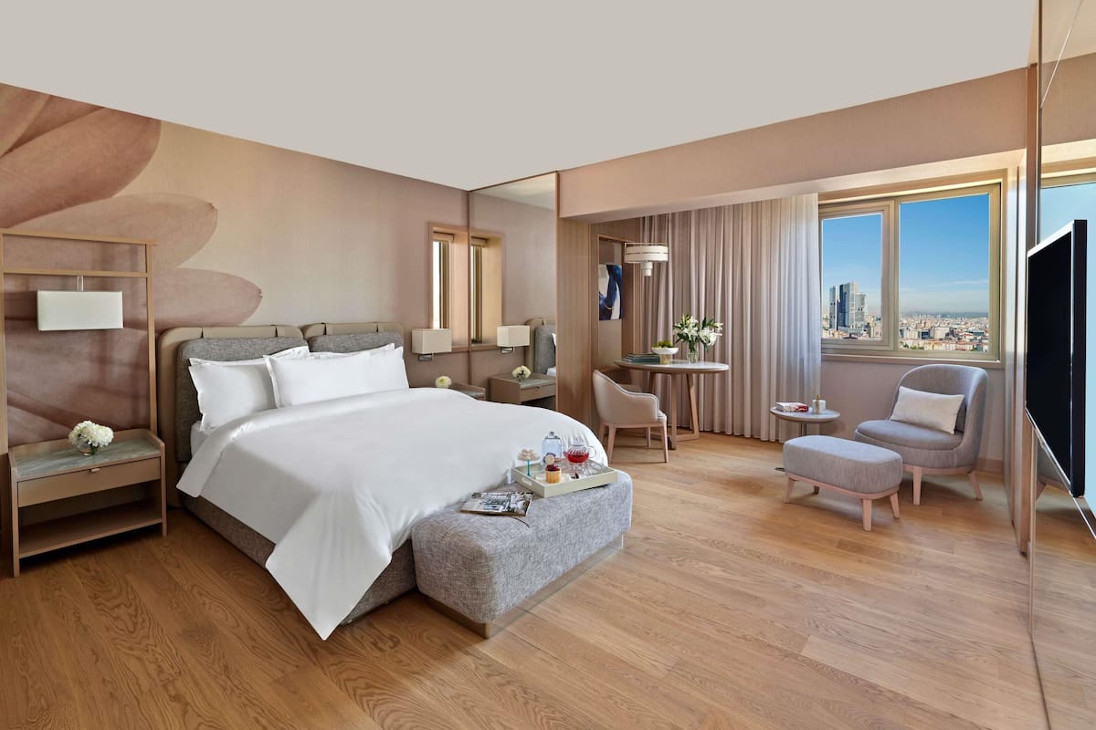 A modern hotel room with a large bed, a desk, a chair, and a view of the city.