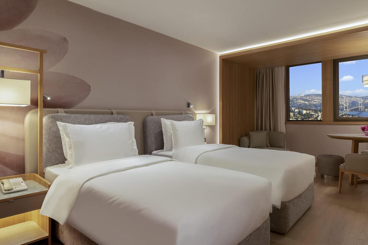 A hotel room with two beds, a small table, a chair, a lamp, and a window with a city view.