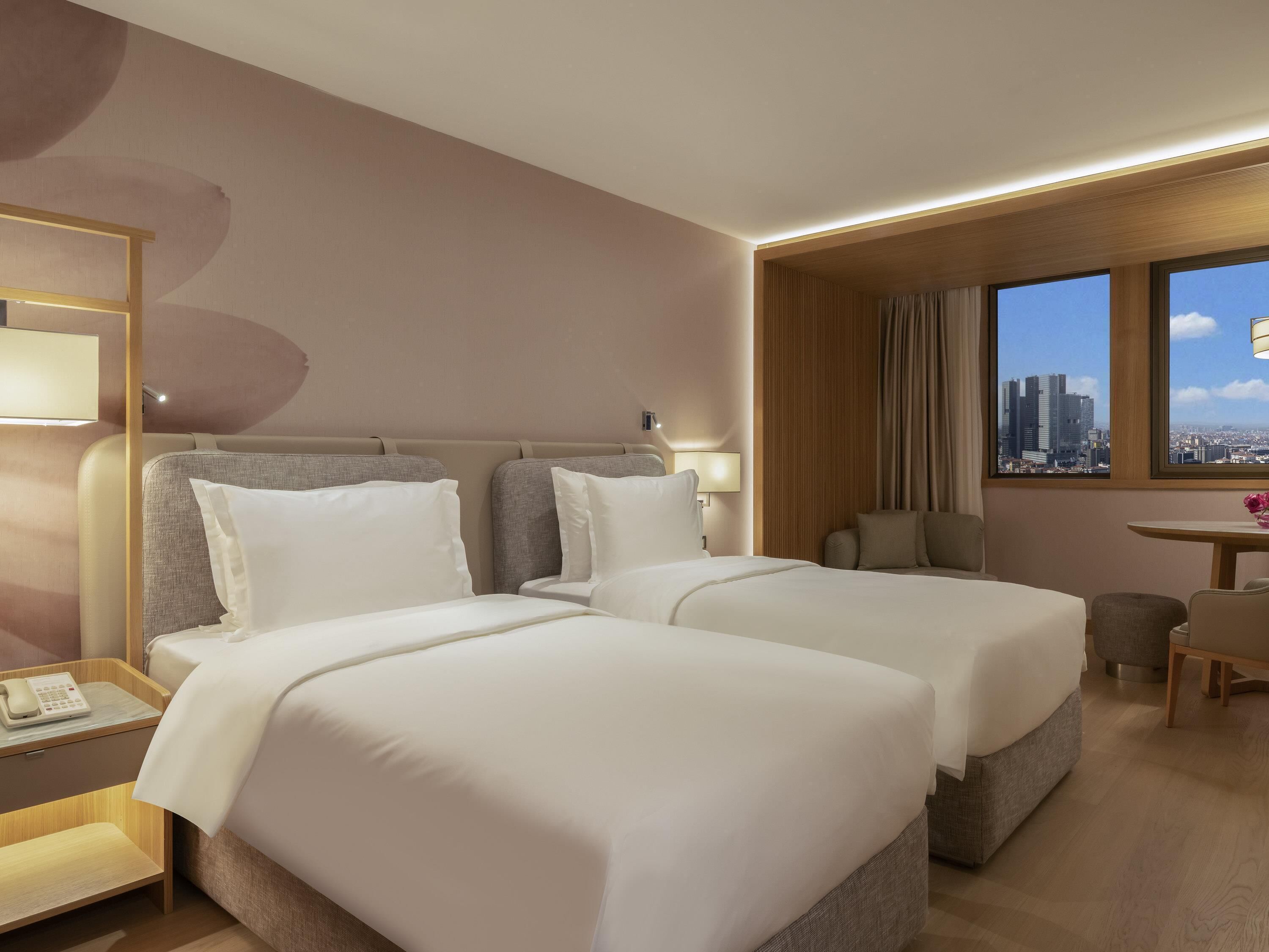 Ein Hotelzimmer mit zwei Betten, einem Schreibtisch, einem Stuhl, einer Lampe und einem Fenster mit Blick auf die Stadt.