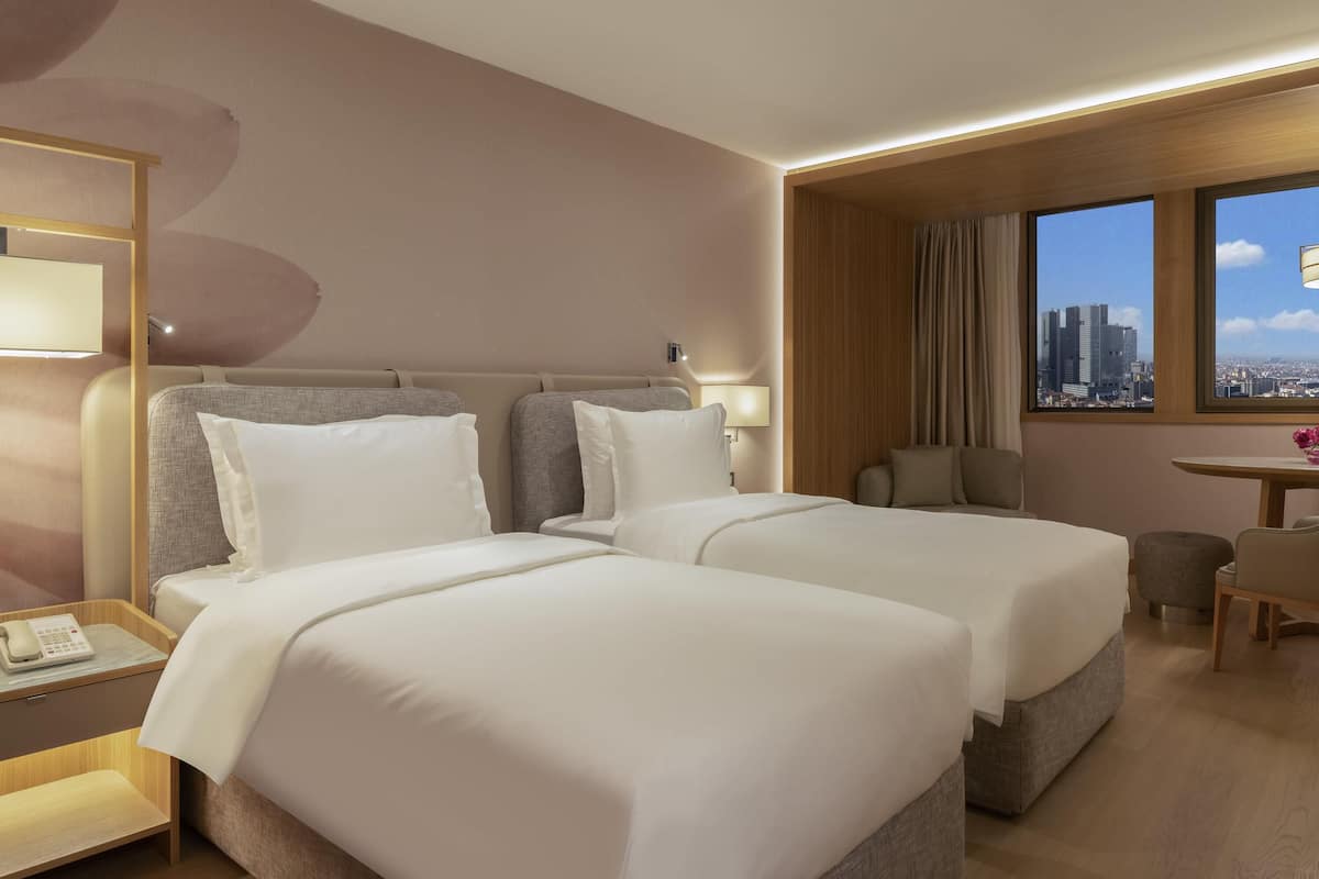 Ein Hotelzimmer mit zwei Betten, einem Schreibtisch, einem Stuhl, einer Lampe und einem Fenster mit Blick auf die Stadt.