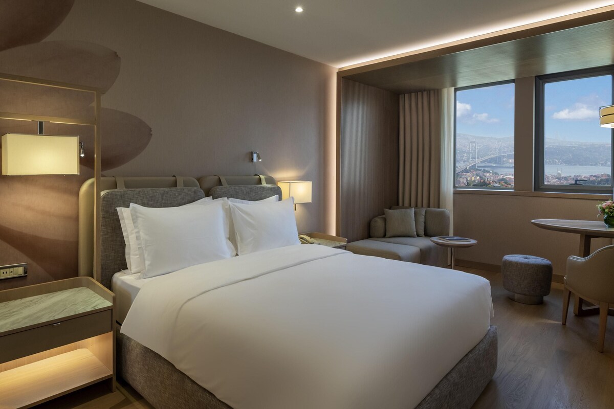 Habitación de hotel con una cama grande, un escritorio, una silla, una mesita y vistas a la ciudad por la ventana.
