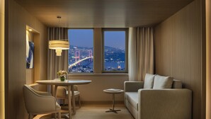 A modern hotel room with a dining area, a sofa, and a view of a cityscape with a bridge. - Mövenpick Hotel Istanbul Bosphorus (Istanbul)