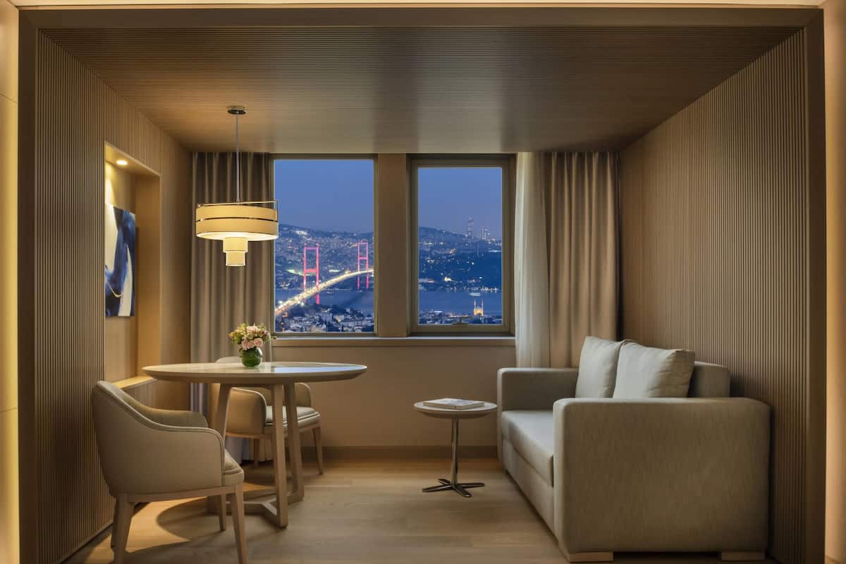 A modern hotel room with a dining area, a sofa, and a view of a cityscape with a bridge.