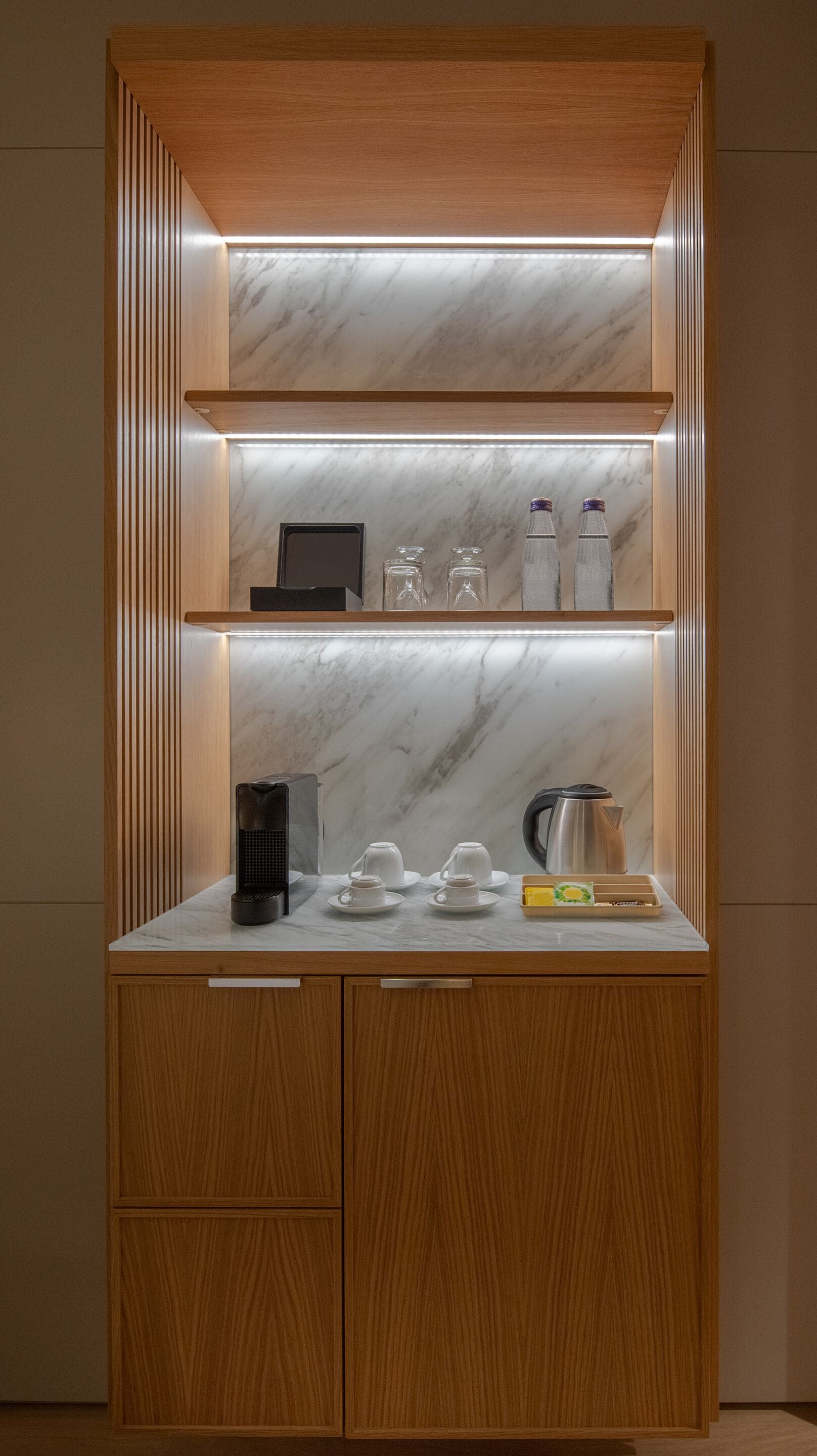 A wooden cabinet with a marble countertop, shelves with glassware, and a coffee maker.