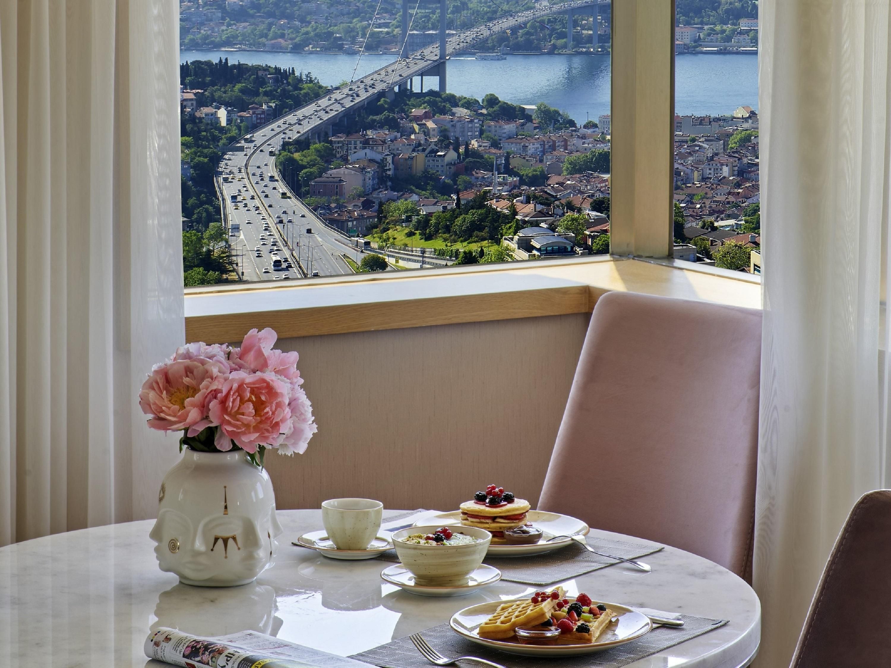 A dining table with a vase of flowers, plates of food, and a view of a bridge and cityscape through a window.