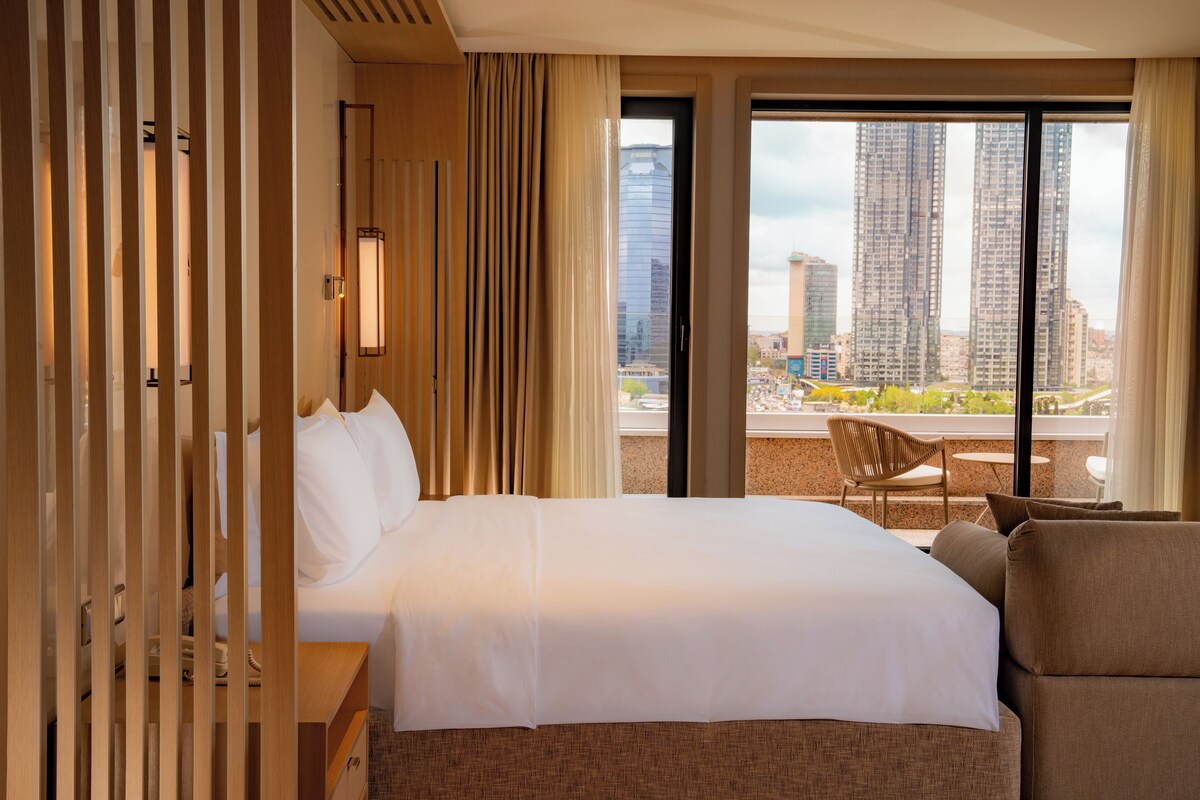 A hotel room with a large bed, a view of tall buildings, and a balcony with a chair.