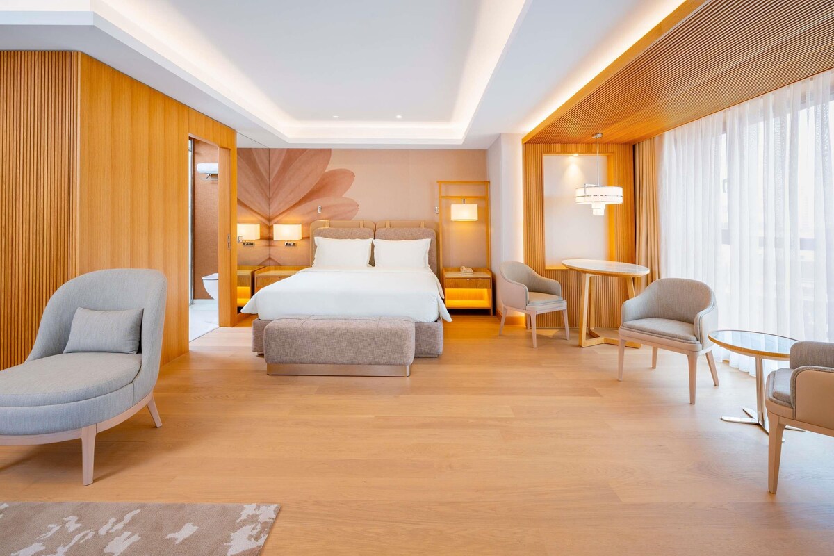 Un hotel moderno con una cama grande, dos sillones, una mesita y vistas de la ciudad a través de amplios ventanales.