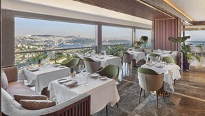 A restaurant with tables set for dining, large windows offering a city view, and a modern interior design. - Mövenpick Hotel Istanbul Bosphorus (Istanbul)