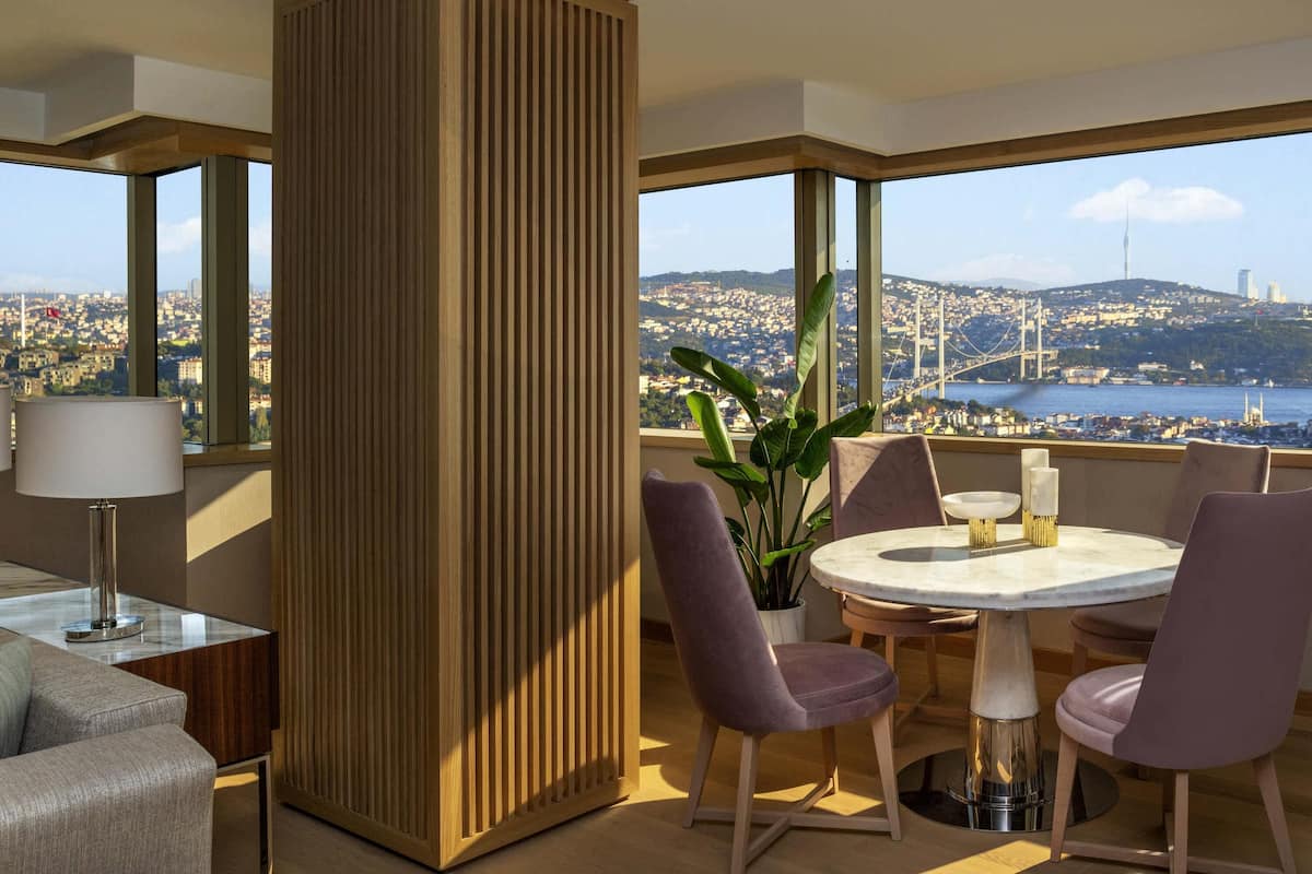 A modern hotel room with a large window offering a city view, a round table with chairs, and a tall wooden partition.