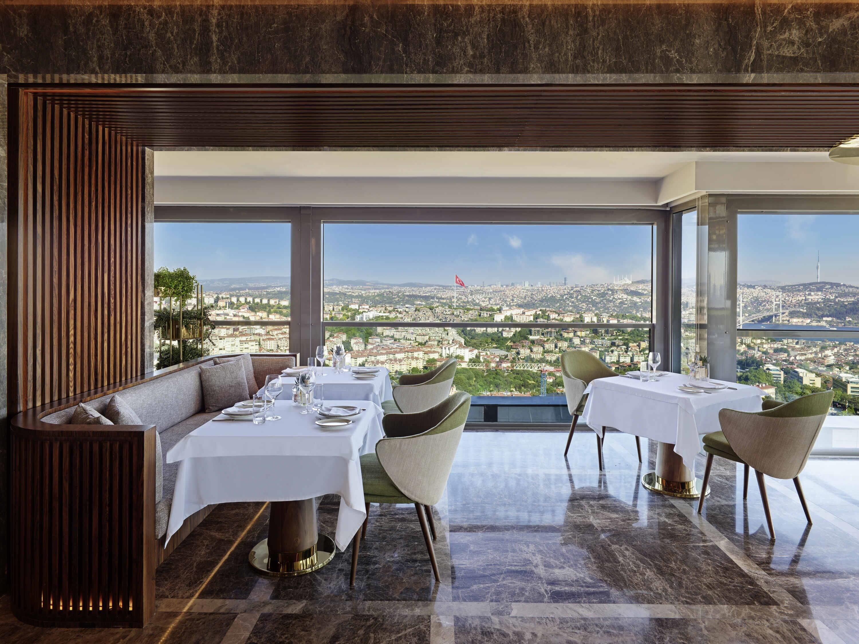 Un restaurante con vistas elevadas a un paisaje urbano, con mesas dispuestas con manteles blancos y sillas.