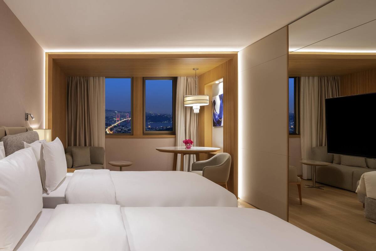 A hotel room with a large bed, a desk, a chair, a TV, and a view of a cityscape.