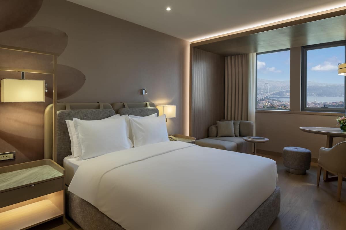 Ein Hotelzimmer mit einem großen Bett, einem Schreibtisch, einem Sessel, einem kleinen Tisch und Blick auf eine Stadtlandschaft durch das Fenster.