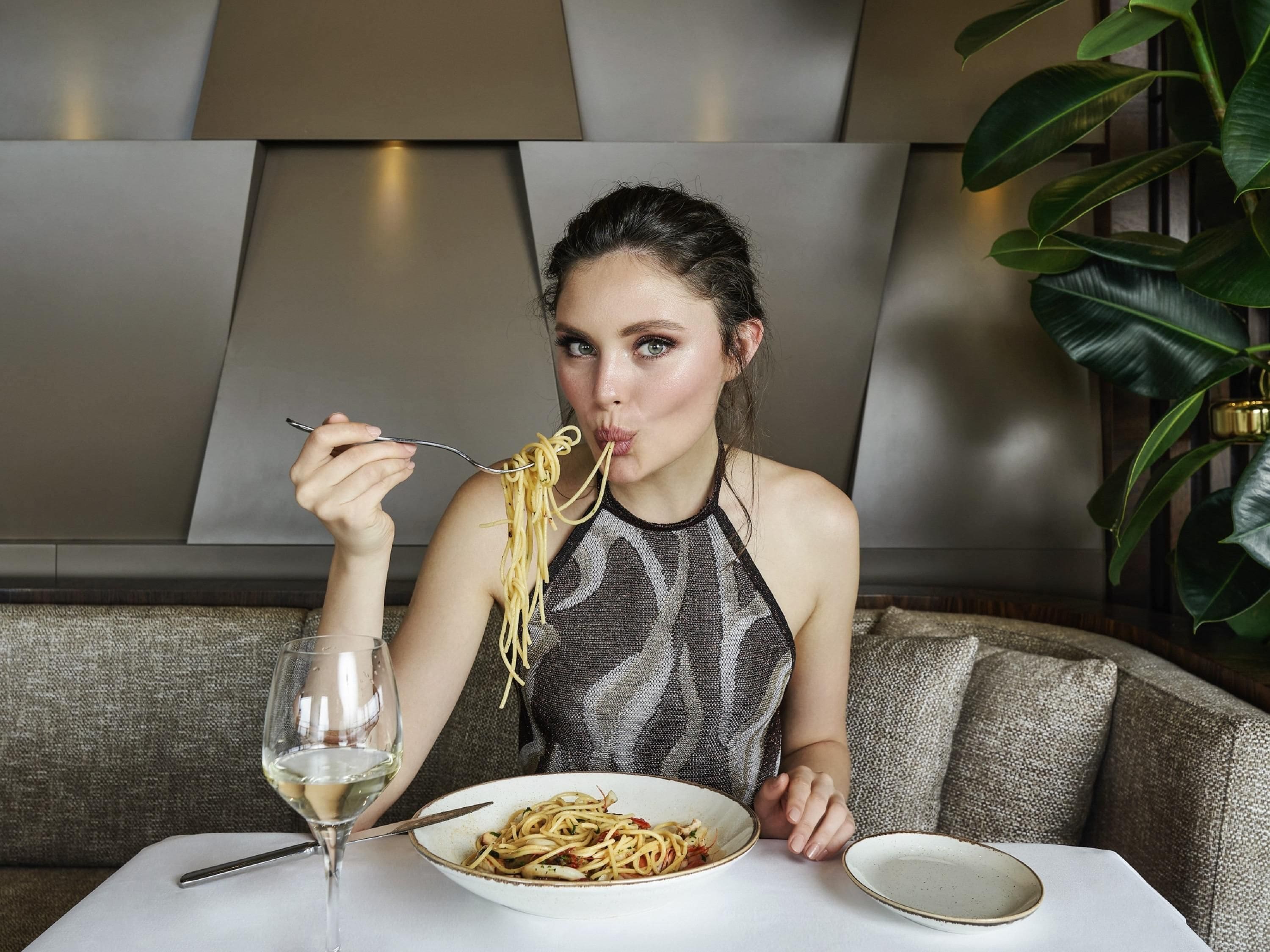 A person seated at a table with a plate of spaghetti, a glass of white wine, and a bowl. The setting includes a sofa and a plant.