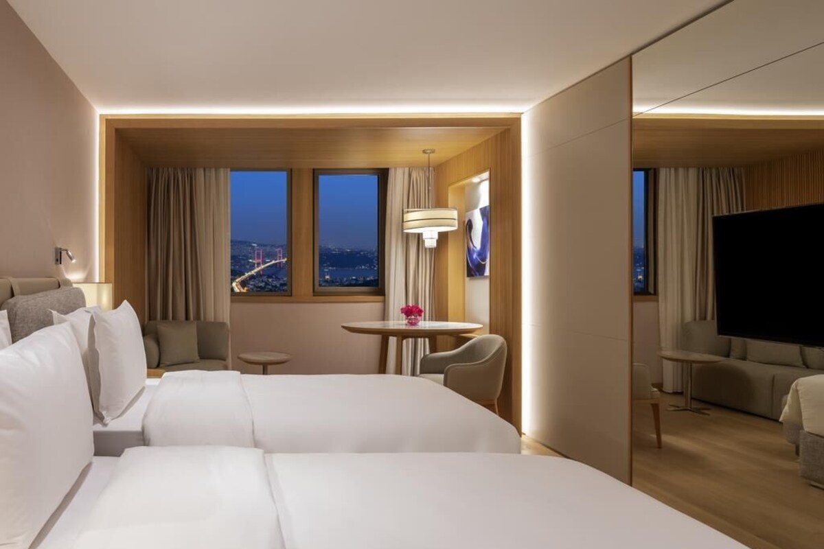 A hotel room with two beds, a TV, a small table with a lamp, and a view of the cityscape.