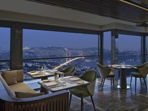 A modern dining area with a table set for two, large windows offering a cityscape view, and a high ceiling with a wooden slat design. - Mövenpick Hotel Istanbul Bosphorus (Istanbul)