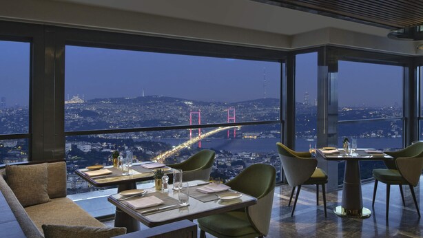 A modern dining area with a table set for two, large windows offering a cityscape view, and a high ceiling with a wooden slat design. - Mövenpick Hotel Istanbul Bosphorus (Istanbul)