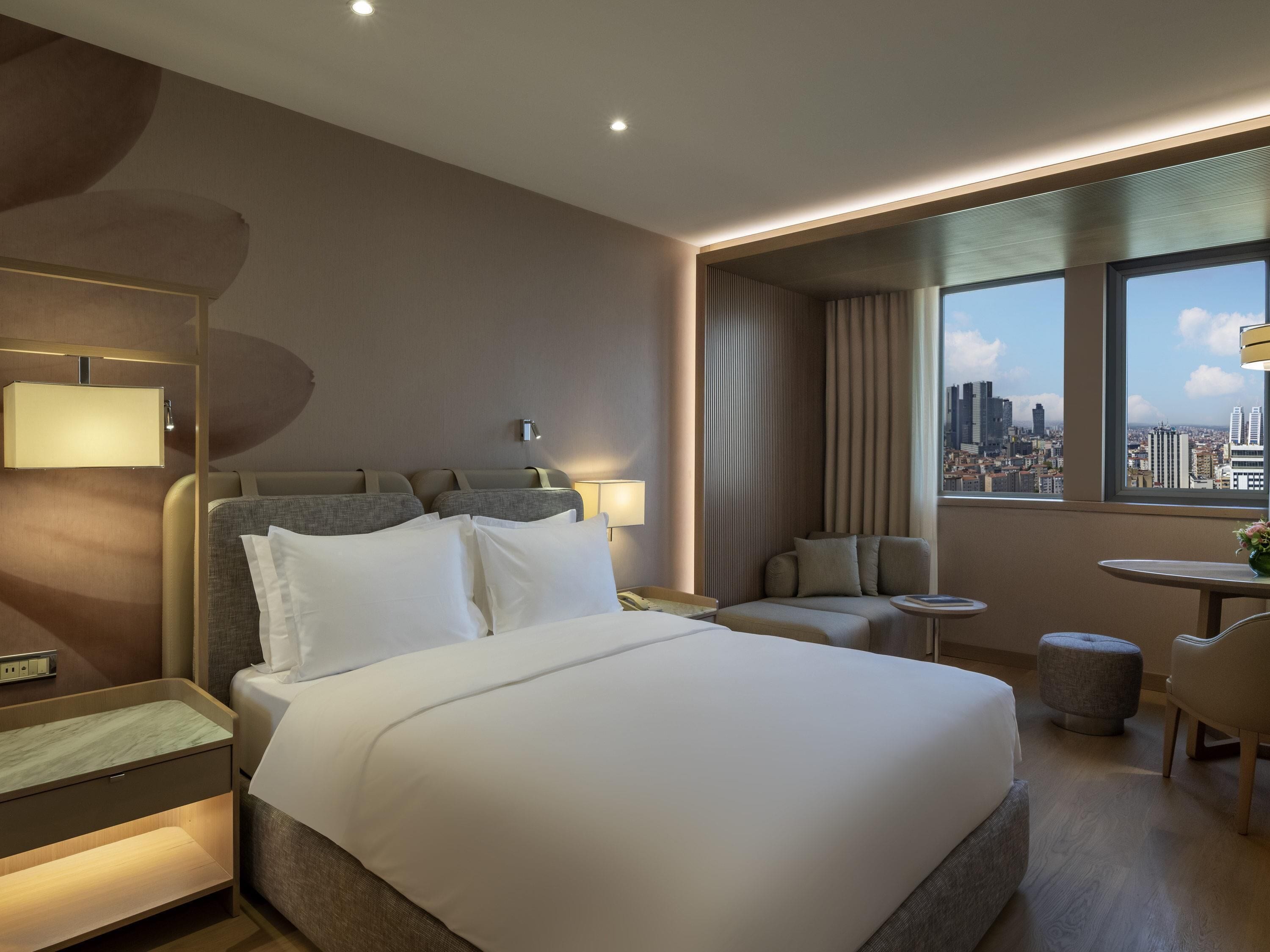Ein Hotelzimmer mit einem großen Bett, einem Schreibtisch, einem Sessel, einem kleinen Tisch und Blick auf die Stadt.