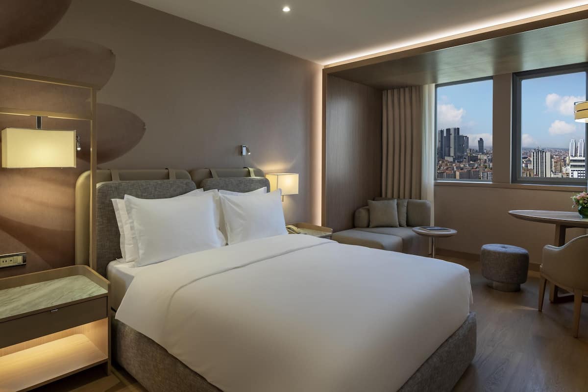 Ein Hotelzimmer mit einem großen Bett, einem Schreibtisch, einem Sessel, einem kleinen Tisch und Blick auf die Stadt.