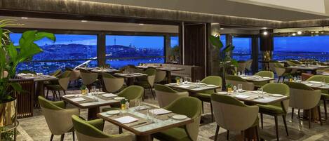 Un restaurante con mesas y sillas dispuestas para comer, grandes ventanales que ofrecen vista a la ciudad, y una decoración interior moderna.