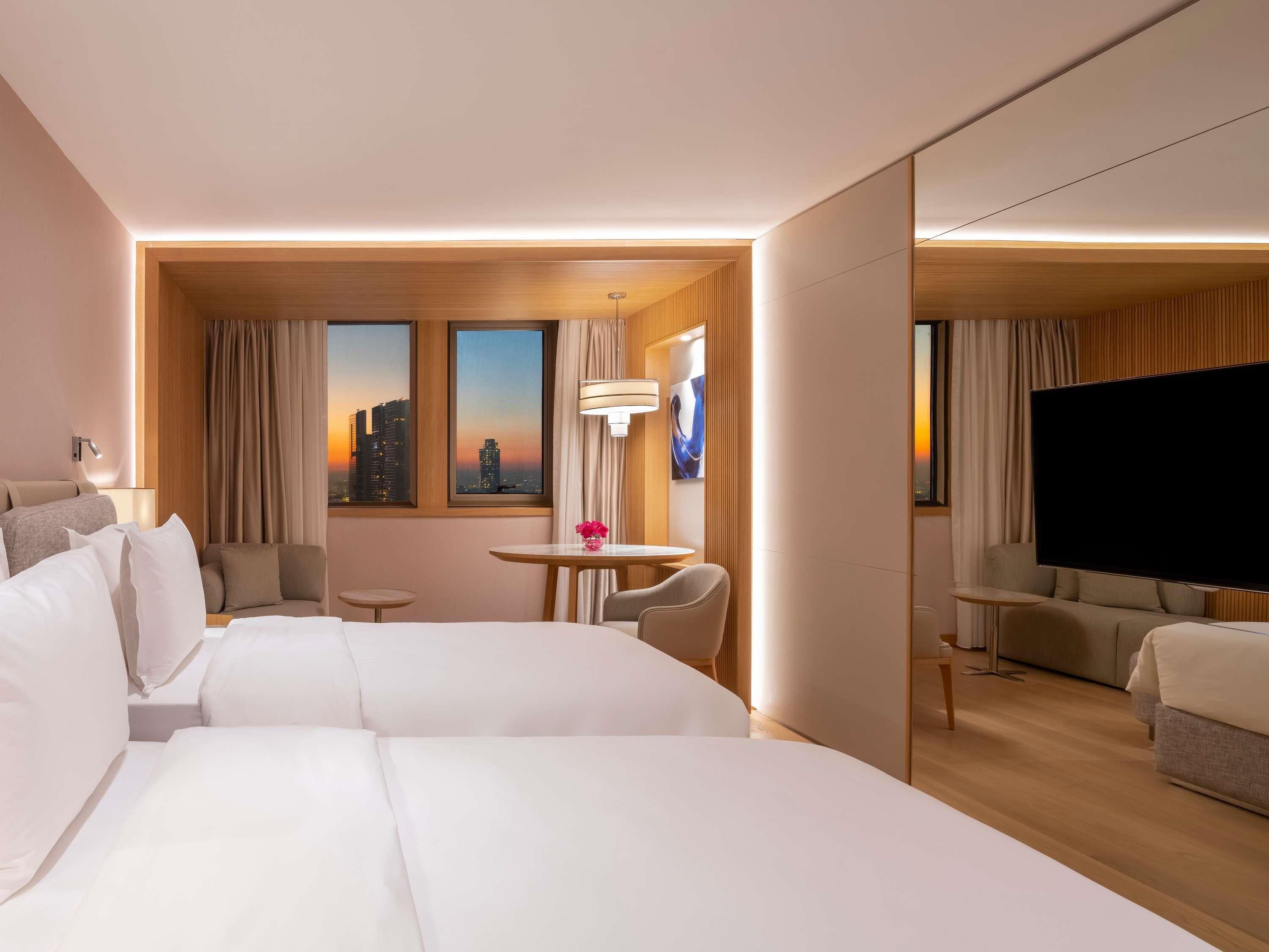 Ein Hotelzimmer mit zwei Betten, einem Fernseher, einem kleinen Tisch mit einer Vase und Blick auf die Stadt.