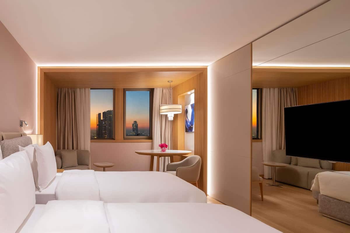 Ein Hotelzimmer mit zwei Betten, einem Fernseher, einem kleinen Tisch mit einer Vase und Blick auf die Stadt.